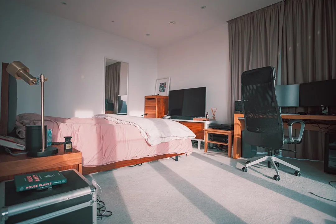 Bedroom with desk setup, ergonomic chair, dual monitors, and pink bedding showing work-sleep space combination
