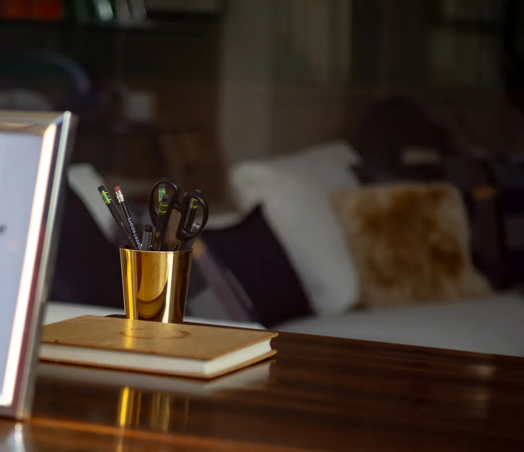 Gold pencil cup with scissors and pencils on notebook atop wooden desk with blurred couch and pillows in background